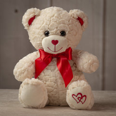 Cream colored stuffed bear with red bow on neck and embroidered hearts on left paw