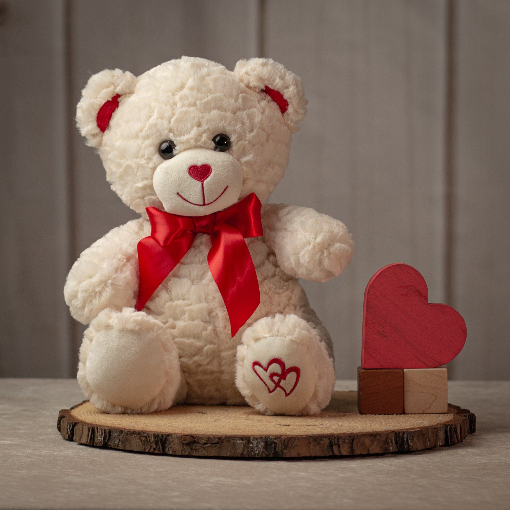 Cream colored stuffed bear with red bow on neck and embroidered hearts on left paw