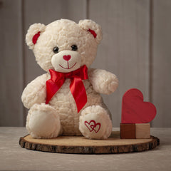 Cream colored stuffed bear with red bow on neck and embroidered hearts on left paw
