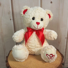 Cream colored stuffed bear with red bow on neck and embroidered hearts on left paw
