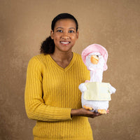 A woman holds a white goose that is 14 inches tall while sitting wearing glasses and a light pink cap holding a nursery rhyme book