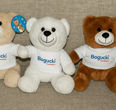 Three plush teddy bears wearing Bogucki Foundation t-shirts sitting side-by-side on a couch