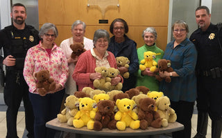Members of the GFWC Herndon Woman’s Club pose with police officers and a table full of teddy bears inside a police department lobby. Everyone is smiling, and the bears are a mix of brown, yellow, and tan.
