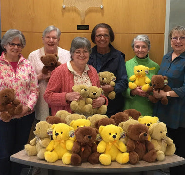Members of the GFWC Herndon Woman’s Club pose with police officers and a table full of teddy bears inside a police department lobby. Everyone is smiling, and the bears are a mix of brown, yellow, and tan.