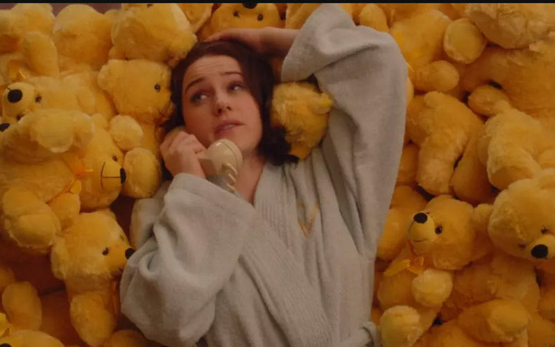 Actress in bathrobe laying on a bed of yellow teddy bears from The Marvelous Mrs. Maisel