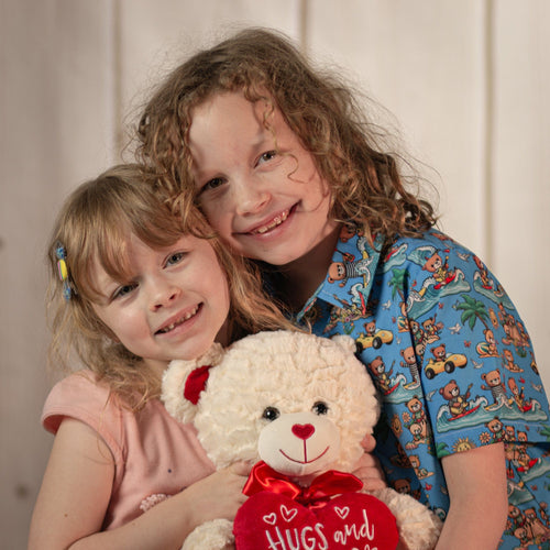 Two little girls holding a cream colored bear with a red bow holding a stuffed heart that says, "Hugs and Kisses."