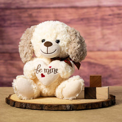 A cream colored dog with brown ears that is 10 inches tall while sitting holding a cream Be Mine heart on top of a piece of wood