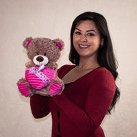 A woman holding a brown bear that is 10 inches tall while sitting holding a plush heart wrapped in a Valentine bow tie
