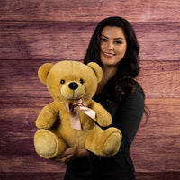 A woman holds a beige bear that is 14 inches tall while sitting wearing a matching bow