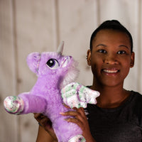 A woman holding a purple unicorn that is 14 inches while laying down with embroidered eyes, shiny paws, wings and horn.