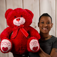A woman holding a red bear that is 19.5 inches tall with a heart nose and embroidered red hearts on its feet