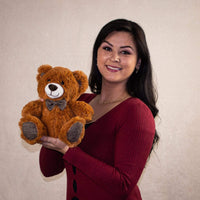A woman holds a brown bear that is 10 inches tall while sitting wearing a bowtie that matches his ears and paw prints