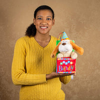 A woman holds a animated/singing birthday dog that pops out of a box that that says "HAPPY Birthday"