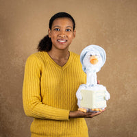 A woman holds a white goose that is 14 inches tall while sitting wearing glasses and a light blue cap holding a nursery rhyme book
