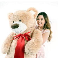 A woman holds a beige bear that is 60 inches tall while standing
