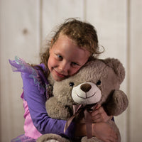 A little girl holding a sitting brown bear that is 14 inches tall while sitting with paw prints on its feet