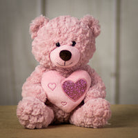 A pink bear that is 10 inches tail while sitting holding a heart accented with smaller shiny hearts