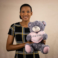 A woman holds a gray bear that is 13.5 inches tall while sitting holding a pink Love You heart