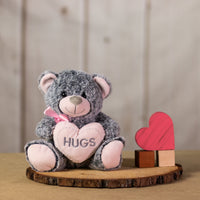 A gray bear that is 9 inches tall while sitting holding a pink heart that says "HUGS" sitting next to wooden blocks