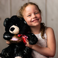 A little girl holding a black bear that is 17 inches tall with paw prints on its feet wearing a red heart covered bow holding a shiny, gold heart