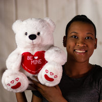 A woman holding a white polar bear that is 14 inches tall while sitting holding a red "BE MINE" heart with embroidered red hearts on its feet