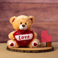 A beige bear that is 10 inches tall while sitting with glittery feet and ears holding a glittery red "LOVE" heart next to wooden blocks
